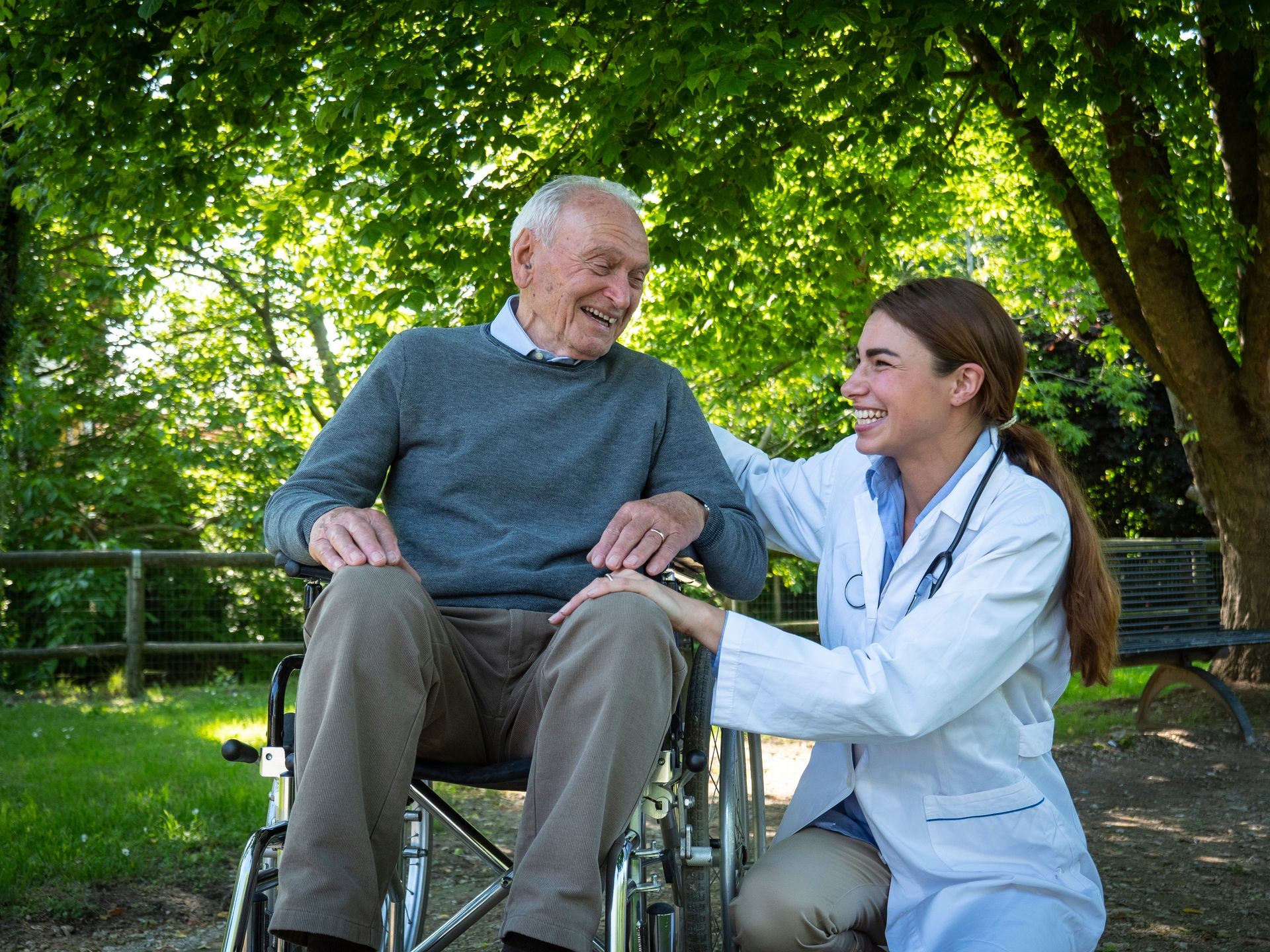 Portrait of carefree and happy young woman social worker or nurse is stroking the hand of senior man in a wheelchair as sign of care and support during they are having fun to talk to each other in a g